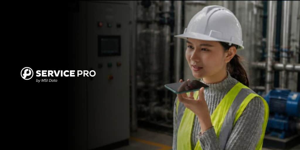 A technician in a hard hat and safety vest using voice AI on her phone in an industrial facility, representing AI-powered field service management software.