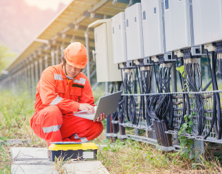 Field technician at a work site, squatting by power & energy equipment and holding a laptop.